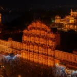 Jaipur city during Diwali 2025 glowing with golden lights — view of Hawa Mahal, Amer Fort, and Jal Mahal beautifully illuminated, showcasing the best places to visit in Jaipur on Diwali. The streets of Johari Bazaar are crowded with people shopping, celebrating, and enjoying the festive atmosphere in the Pink City of India. Best Places to Visit in Jaipur on Diwali 2025